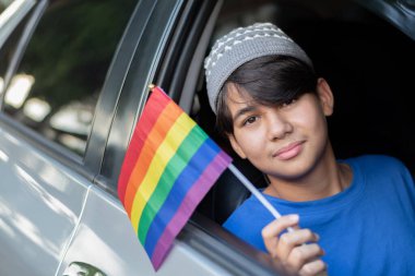 Araba camından gökkuşağı bayraklı bir bayrak tutan gülümseyen genç çocuk, LGBTQ + gurur ve çeşitliliği kutluyor. Özgürlük kavramı, eşitlik, kendini ifade etme ve kapsayıcılık için gençlik desteği.