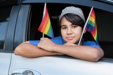 Araba camından gökkuşağı bayraklı bir bayrak tutan gülümseyen genç çocuk, LGBTQ + gurur ve çeşitliliği kutluyor. Özgürlük kavramı, eşitlik, kendini ifade etme ve kapsayıcılık için gençlik desteği.
