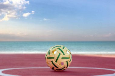 Sepak, kumsal sahasının kum zemininde topu, bulanık mavi gökyüzü ve deniz arka planı, Güneydoğu Asya ülkelerindeki eğlence aktivitesi ve açık hava sporları konsepti.