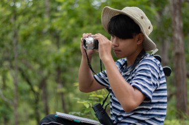 Genç bir kâşif ormanda bir yaprak ve ağaç fotoğraflıyor. Alan çalışması için dürbünle, kamerayla ve defterle doğayı belgeliyor..