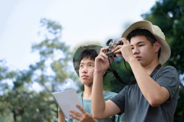Genç bir adam ormanda bir ağaç gövdesine yaslanmış bir kamera tutuyor. Fotoğrafçılığa odaklanmış, doğanın karmaşık detaylarını yakalıyor..