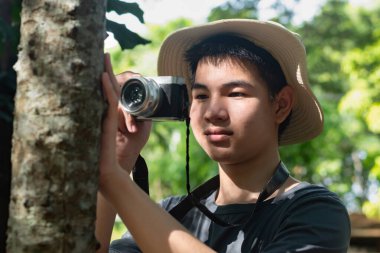 Genç bir adam ormanda bir ağaç gövdesine yaslanmış bir kamera tutuyor. Fotoğrafçılığa odaklanmış, doğanın karmaşık detaylarını yakalıyor..