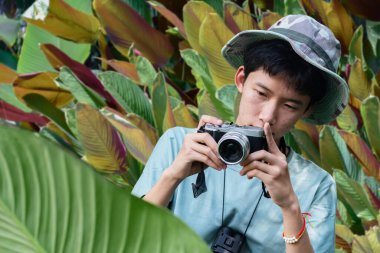 Genç bir adam ormanda bir ağaç gövdesine yaslanmış bir kamera tutuyor. Fotoğrafçılığa odaklanmış, doğanın karmaşık detaylarını yakalıyor..