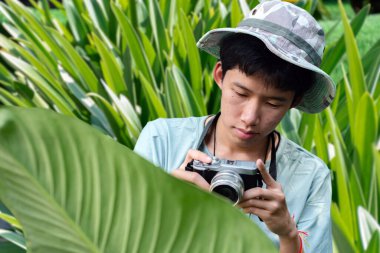 Genç bir adam ormanda bir ağaç gövdesine yaslanmış bir kamera tutuyor. Fotoğrafçılığa odaklanmış, doğanın karmaşık detaylarını yakalıyor..