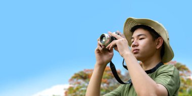 Genç bir adam ormanda bir ağaç gövdesine yaslanmış bir kamera tutuyor. Fotoğrafçılığa odaklanmış, doğanın karmaşık detaylarını yakalıyor..