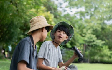 Doğayı keşfeden iki Asyalı çocuk, biri dürbün kullanıyor diğeri de not yazıyor. Yeşil, orman benzeri bir ortamda açık hava öğrenimi, merak ve çocukluk macerası..