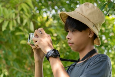 Kova şapkalı, açık havada dijital kamerayla fotoğraf çeken parlak yeşil arka planlı genç bir çocuğun yan görüntüsü..