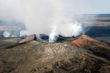Hawaii 'deki Kilauea volkanının krateri, yoğun duman ve buharla yükseliyor. Kraterin engebeli kenarı ve etrafındaki lav alanları sık sık püskürmeleriyle bilinen bu bölgedeki yoğun volkanik aktiviteyi vurguluyor..