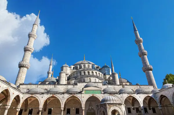 Görüntüde Türkiye 'nin İstanbul kentindeki görkemli Mavi Cami, yüksek minareleri, büyük kubbeleri ve parlak mavi bir gökyüzü altında karmaşık bir şekilde tasarlanmış avlusu yer alıyor.