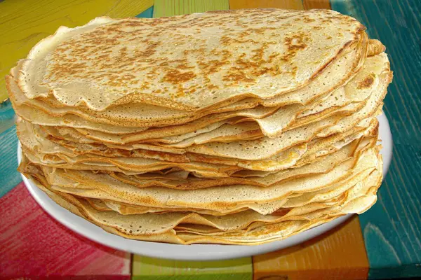 stock image The image showcases a large stack of thin, golden-brown crepes piled high on a white plate. Each crepe is delicately cooked with crispy edges and a soft, slightly airy texture. The vibrant, multicolored wooden table adds a rustic charm to the scene