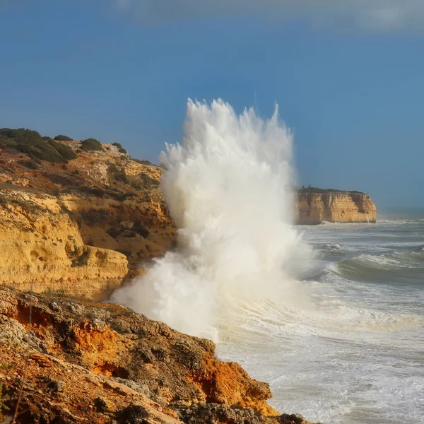 Wave splashing into the air as it crashes into orange cliffs on a windy winter day in Carvoeiro, Portugal.
