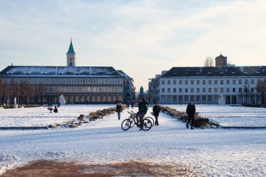 Karlsruhe, Almanya - 12 Şubat 2021: Almanya 'da güneşli bir kış gününde Karlsruhe Sarayı' nın önündeki karlı bir yolda yürüyen ve bisiklete binen insanlar.