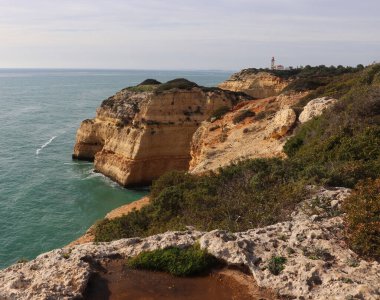 Carvoeiro, Portekiz - 25 Aralık 2019: Arka planda Farol De Alfanzina Deniz Feneri ile Atlantik Okyanusu 'nun yanındaki kireçtaşı uçurumunda kayalar ve çalılar.