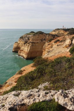 Carvoeiro, Portekiz - 25 Aralık 2019: Güney Portekiz 'de güneşli bir kış gününde Atlantik Okyanusu manzaralı arka planda Farol De Alfanzina Deniz feneriyle kayalıkları ve çalıları olan uçurum.