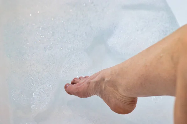 Foot with toes pointed toward water with bubbles in a white bathtub.