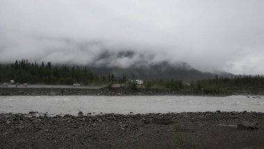 McCarthy, Alaska - July 13, 2023: Campers and cars next to a river with clouds in front of a mountain in the distance on a summer day in Wrangell - St Elias National Park Preserve in Alaska. clipart