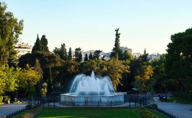 Atina, Yunanistan - 21 Eylül 2024: Yunanistan 'da bir sonbahar öğleden sonrasında parkta bir fıskiyenin arkasındaki ağaçlarda güneş parlıyor.