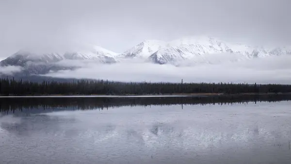 Kanada 'daki Alaska Otoyolu' nda bulutlu bir sonbahar gününde, gölün arkasındaki karlı dağda aydınlık. 