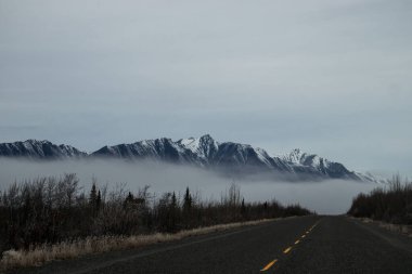 Kanada 'da bulutlu bir sonbahar gününde Alaska otoyolunun arkasındaki dağın eteğinde yoğun sis var.. 