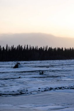 Donmuş nehirde kar, arka planda ağaçlar ve dağlar. Alaska 'nın iç kesimlerinde kış gündönümünde.. 