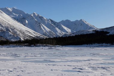 Güneşli bir kış gününde, Alaska 'daki Richardson Otoyolu' nda donmuş nehrin arkasındaki dağın tepesinde kar esiyor.. 