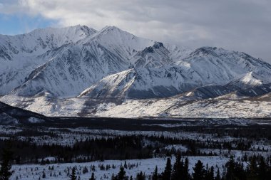 Karlı dağların eteklerinde parlayan güneş bulutlu bir kış gününde Alaska 'daki Richardson Otoyolu' nda.. 