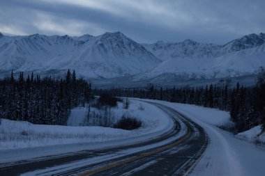 Alaska otoyolunun arkasındaki karlı dağlar karanlık, bulutlu bir kış günü Tok yakınlarında Alaska 'nın iç kesimlerinde. 