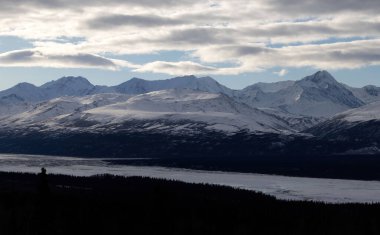 Alaska 'nın iç kesimlerindeki Richardson Karayolu' nda bulutlu bir kış gününde bir nehrin üzerindeki dağların üzerindeki ışık havuzları..