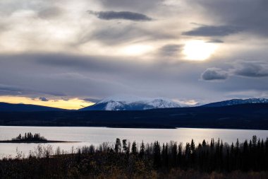 Alaska otobanı boyunca uzanan bir gölün arkasındaki bulutların üzerinde parlayan güneş Kanada 'da karanlık bir sonbahar öğleden sonrasında. 