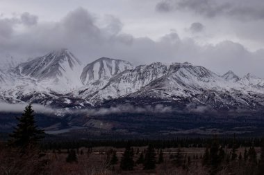Kanada 'da soğuk, karanlık bir sonbahar gününde Alaska otobanındaki karlı dağların üzerinde bulutlar..