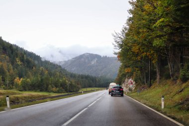 Bavyera, Almanya - 6 Ekim 2019: Almanya 'nın Bavyera şehrinde yağmurlu bir sonbahar gününde, yoldaki arabaların yanında ağaçlar ve arka planda bir dağ. 