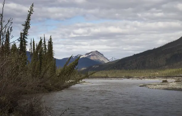 Alaska 'da bulutlu bir bahar gününde Dalton Otoyolu' ndaki bir nehrin arkasındaki dağın tepesinde güneş parlıyor..