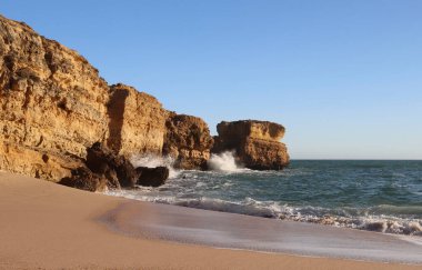 Algarve, Portekiz 'de güneşli bir günde kumlu bir plajın yanındaki kayalıklara su sıçrıyor.. 