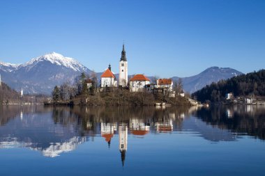 Bled, Slovenya - 16 Ocak 2022: Bled Island 'daki kilise, Slovenya' da güneşli bir kış gününde arka planda dağlarla birlikte sudaki yansımayla.