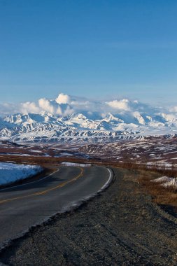 Alaska 'da güneşli bir bahar gününde, beyaz bulutlarla kaplı, karlı dağlara uzanan Denali Otoyolu' nun Paved bölümü..