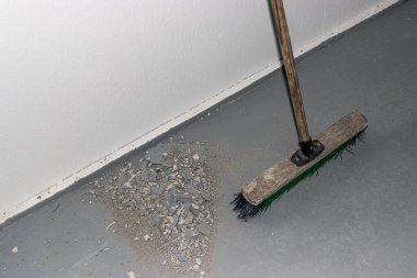 Gri bir zemindeki enkazı süpüren bir süpürge görüntüsü, basit, çıplak bir iç çevre içindeki temizlik ve temizlik faaliyetlerini vurguluyor.