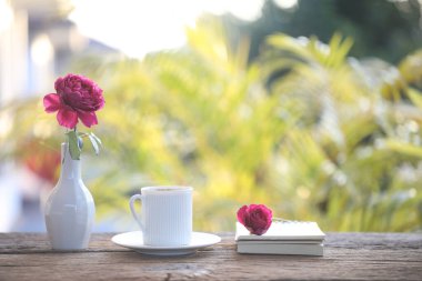 Red rose in white vase and white coffee cup and notebook on wooden table