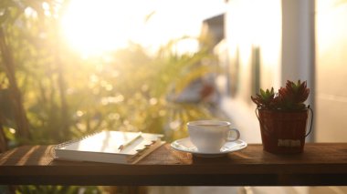 Kahve fincanı, defter ve güneş ışığı altında ahşap masaya demlik.