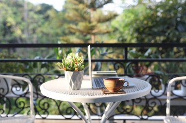Dışarıdaki ahşap çalışma masasının üzerinde çay fincanı. Laptopun yan görüntüsü ve dışarıdaki demlik balkonu.