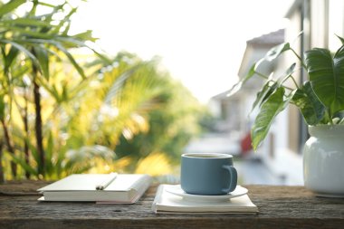 Ahşap tepside mavi kahve fincanı, defter ve Philodendron bitkisi güneş ışığı altında ahşap masada.