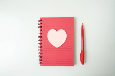 kırmızı defter, kalp notu ve kırmızı kalem beyaz masada.
