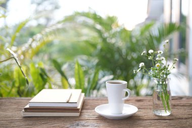 Tahta tepside uzun beyaz kahve fincanı ve bahçede güneş ışığı altında ahşap masa üzerinde bitki içeren defter.