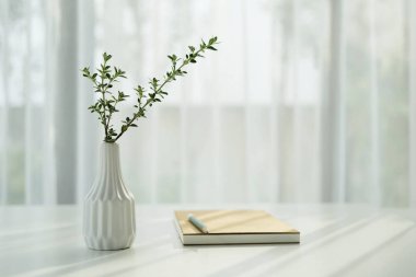 Pencere kenarındaki not defteriyle birlikte beyaz masadaki uzun bitki vazosu, asgari iç mekan, rahat çalışma alanı