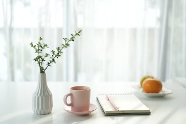 Pencere kenarında not defteri olan uzun bitki vazosu ve pembe bardak. İç mekan, rahat çalışma alanı.
