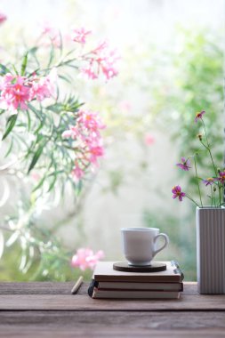 Penceresinde pembe çiçekler ve defter olan beyaz kahve fincanı.