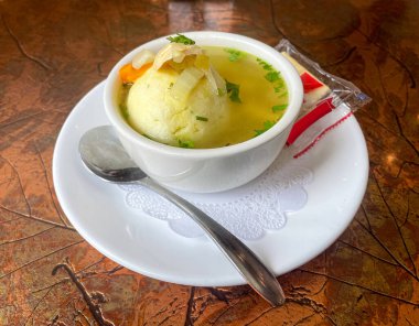 A bowl of matzo ball soup sits on a white plate with a spoon and a pack of saltine crackers. The soup is garnished with chopped vegetables and herbs, served in a clear broth on a textured metal table. clipart