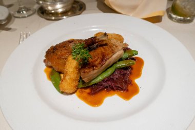 Kurutulmuş siyah biberli ördek göğsü parçaları, lezzetli bir jöle ile parıldıyor, beyaz bir tabağın üzerinde sanatsal bir şekilde oturuyor zarif sofra takımı ve sıcak bir ortam ile çevrili beyaz bir masa örtüsü..