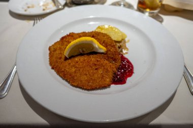 Limon dilimi ile servis edilen altın-kahverengi bir pirzola, keskin kırmızı kızılcık sosu ve beyaz bir tabakta zarif bir şekilde sunulan küçük bir porsiyon patates ve lahana..