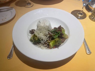 Lezzetli bir biftek ve brokoli tabağı, haşlanmış beyaz pirinç, çıtır pirinç eriştesi ve yeşil soğanla süslenmiş, canlı renklerle beyaz bir tabakta sunuluyor, ve temiz, zarif bir sunum..