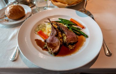 Soslu, kızarmış sebzeli kuzu pirzola, yeşil fasulye, patates püresi ve kremalı soslu gurme tabağı, beyaz bir tabakta ekmek ve resmi bir masada içki ile servis ediliyor.